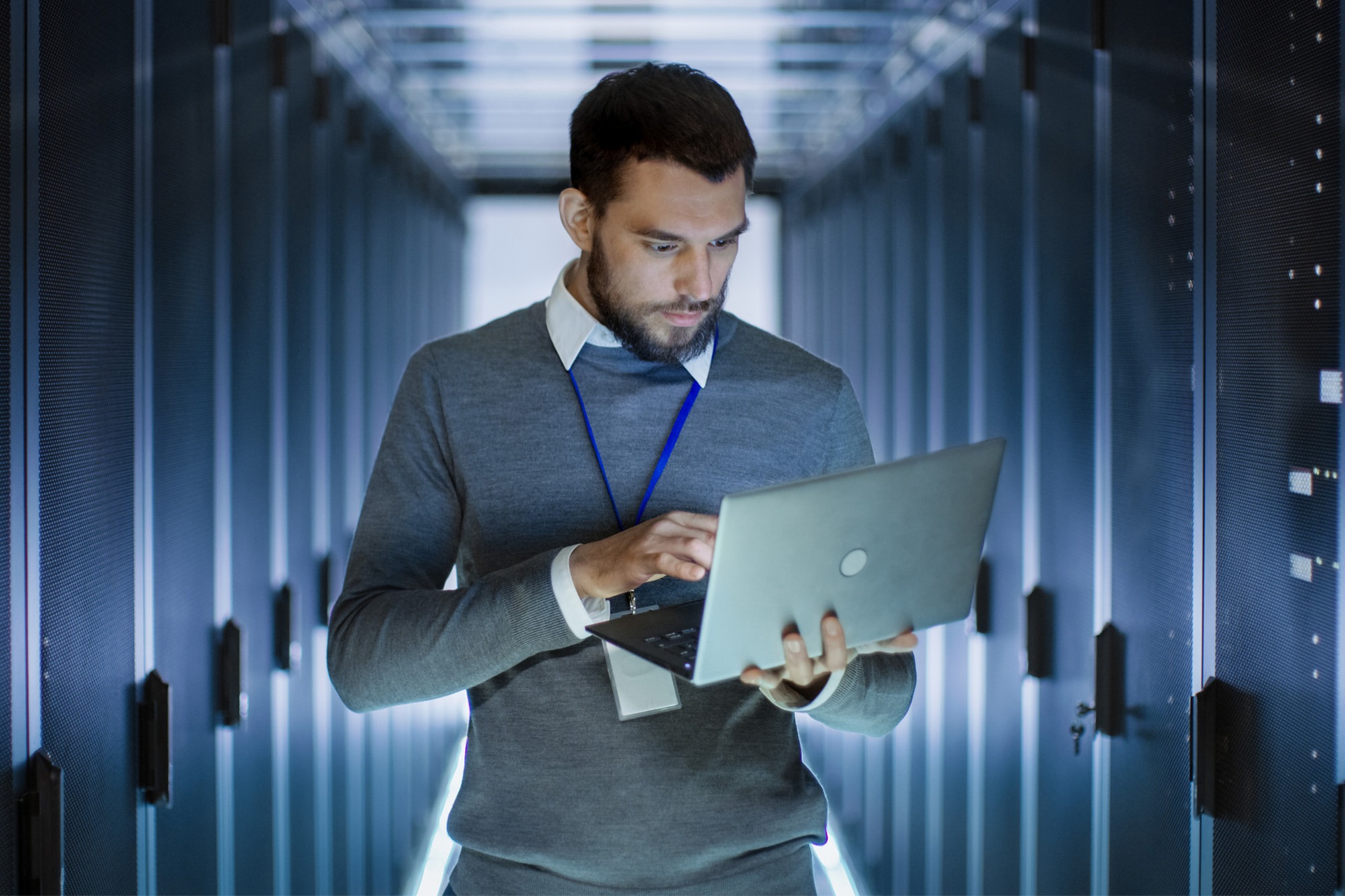 Male Server Engineer Works on a Laptop in Large Data Center.