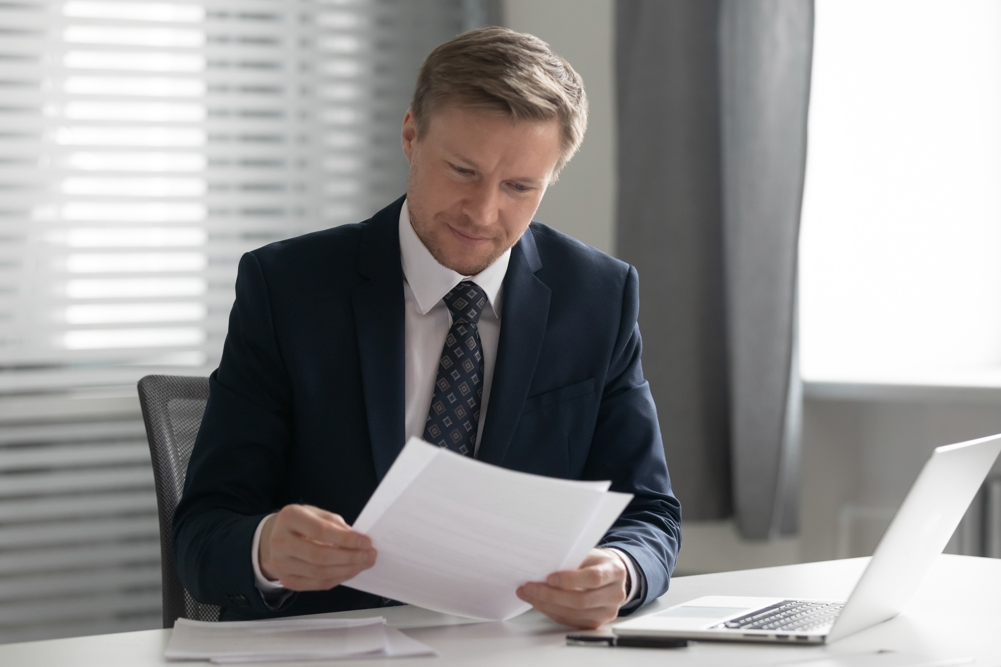 Mid Aged Business Man in Suit Holding Read Document Paper Report Sit at Office Desk, Company CEO Doing Paperwork Feel Satisfied With Good Work Result Receive Loan Approval Salary Payment Tax Refund.