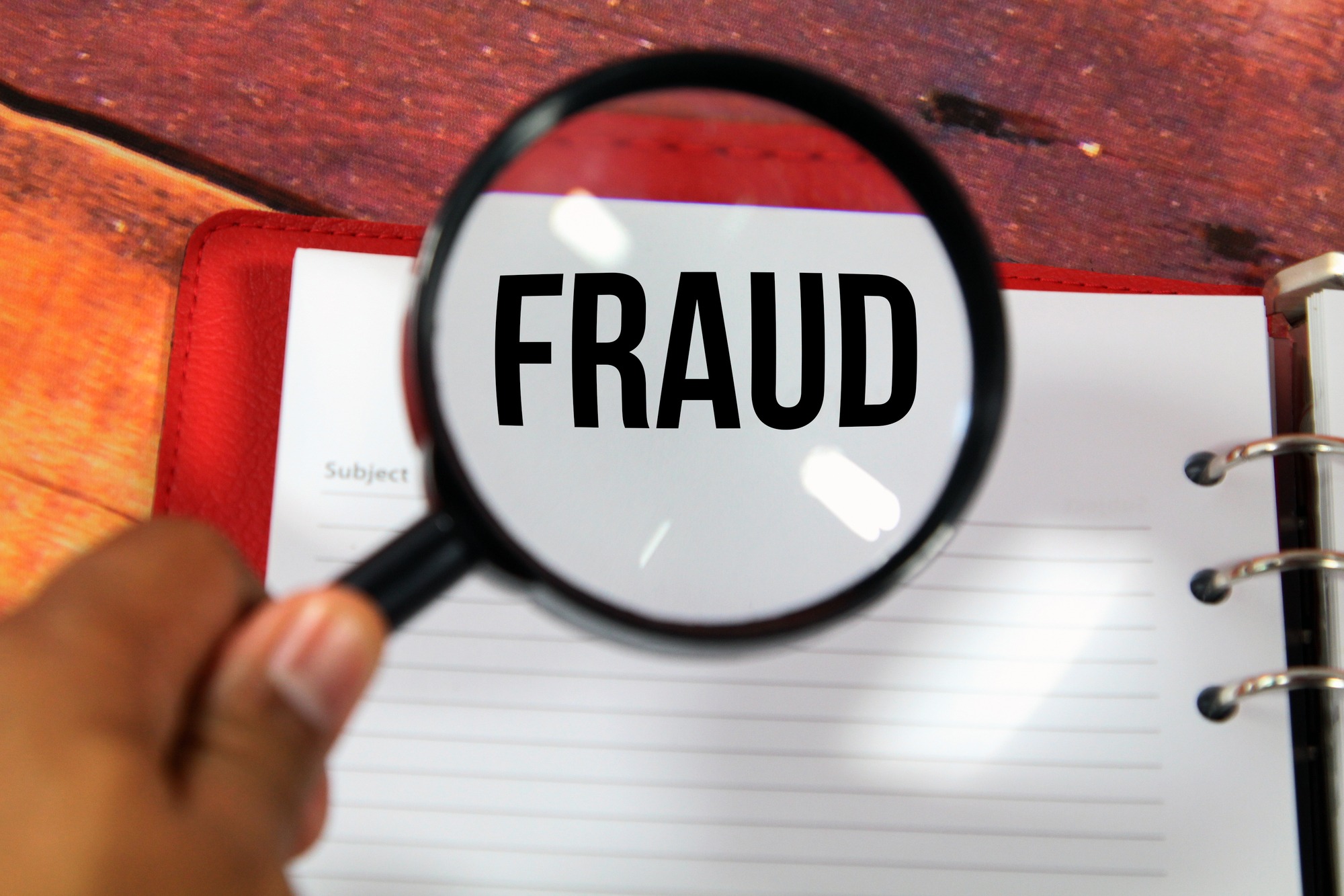 A Concept Image of a Magnifying Glass Zoom on a Notebook Over a Wood Background With a Word Fraud Zoom Inside the Glass.